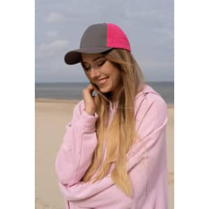 A cap with a grey and pink design is worn by a woman smiling on a beach. The background features sand and sea.