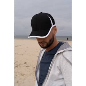 A black cap with white trim, worn by a person at a beach, with a cloudy sky in the background.