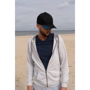 A man wearing a black cap with a blue peak, grey zip-up hoodie, and navy t-shirt stands on a beach.