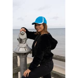 A woman wearing a blue and white cap poses beside a viewing telescope by the beach. The ocean is in the background.