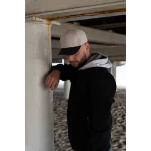 A man leans against a concrete pillar wearing a black jacket and a beige and black cap.