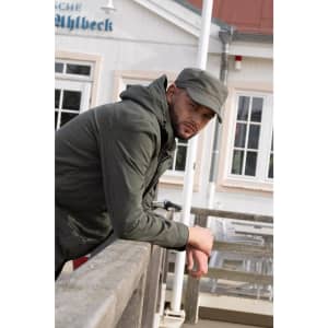 A man wearing an olive green Sports Twill Military Cap leans against a railing, dressed in a jacket. The background features a building.