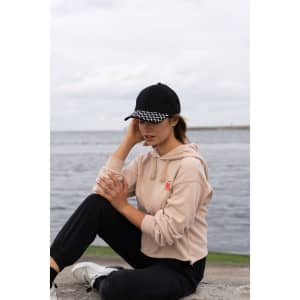 A black cap with a checker plate peak, worn by a woman in a beige hoodie, with a scenic water background.