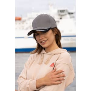 A grey cap with a peak trim, worn by a model in a light-coloured hoodie, set against a maritime backdrop.