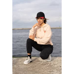 A woman crouches by the water, wearing a beige hoodie, black pants, and a black cap with red inserts.
