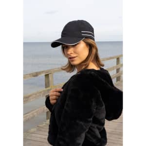 A black microfibre sports cap with white piping, worn by a woman by the seaside. The setting includes a wooden railing.