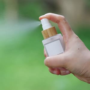 A frosted glass spray bottle with a bamboo cap, being sprayed, set against a green background.