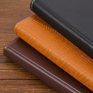 Three diaries stacked: one black, one orange with a textured pattern, and one brown, all featuring a subtle logo.