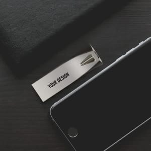 A silver 32 GB USB flash drive rests on a dark wooden surface next to a black smartphone and a black notebook.