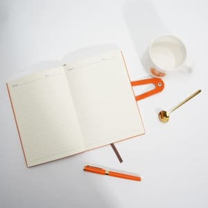 A notebook with cream pages, a ceramic cup, a gold spoon, and an orange pen on a white surface.