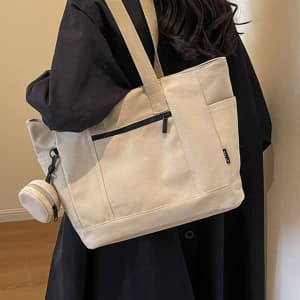 A large capacity canvas shoulder bag in cream, featuring a black zip pocket and a detachable drink bottle holder.