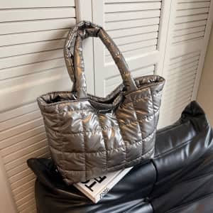 A large quilted tote bag in a shiny silver colour, featuring two handles, resting on a black sofa.