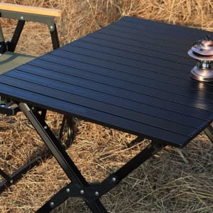 A portable folding camping table in black alongside a chair and a lantern on a grassy surface.