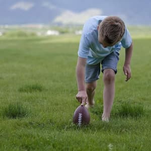 A boy in a blue shirt and shorts kneels on grass, preparing to pick up a brown rugby football.