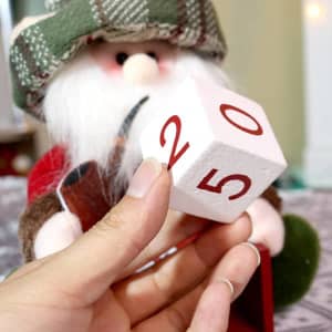 A hand holds a large white block with red numbers, while a plush Santa figure sits nearby, wearing a green hat.
