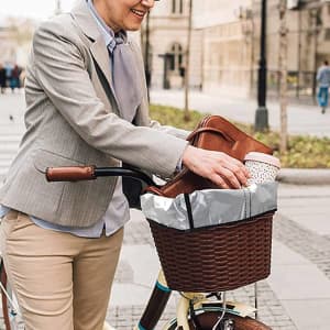 A waterproof bike basket liner in grey is being placed in a woven bike basket. A drink bottle and bag are visible.