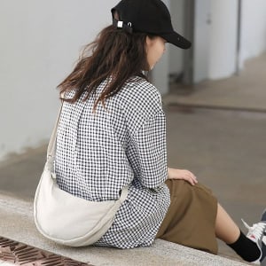 A person wearing a checkered shirt and skirt sits, showcasing a light-coloured personalized crossbody bag.