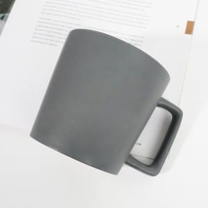 A matte grey stoneware mug with a square handle, resting on a white surface beside a book.