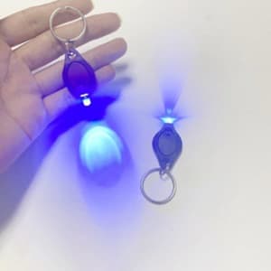 A hand holds two mini LED keychain flashlights, one glowing blue, featuring a silver keyring attachment.