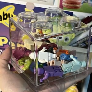 A transparent piggy bank designed like a building block, filled with multi-coloured toy figures.