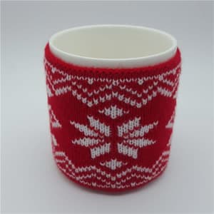 A knitted cup sleeve in red and white with a snowflake design wraps around a plain white mug.