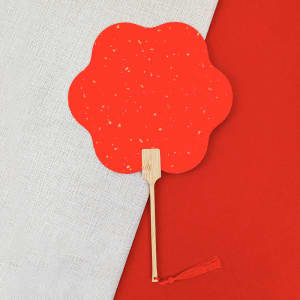 A red bamboo hand fan with a flower-shaped design, featuring a tassel on the handle, against a textured background.