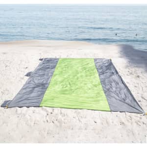 A waterproof beach picnic blanket in grey and bright green laid out on sandy beach near water.