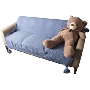 A blue knitted throw blanket with tassels draped over a sofa, alongside a large brown teddy bear.