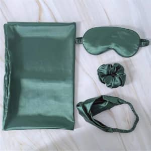A green satin pillowcase, eye mask, hairband, and scrunchie set arranged on a marble surface.