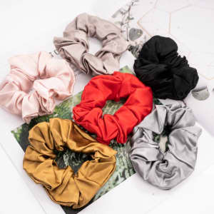 A set of six silk hair scrunchies in pink, beige, red, black, gold, and silver displayed on a flat surface.