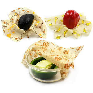 Reusable beeswax wraps in cream with banana patterns, covering an avocado, a red bell pepper, and a bowl with green food.