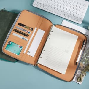 A5 padfolio with a zipper, featuring a tan interior, card slots, a notepad, and a pen. It has a branding logo.