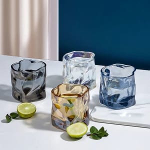A set of four colourful irregular whiskey cups in blue, clear, amber, and dark blue, accompanied by lime halves and mint leaves.