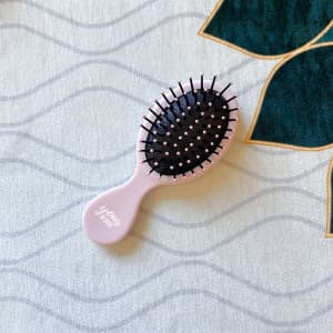 A pink air cushion comb with black bristles sits on a textured background featuring green leaves.