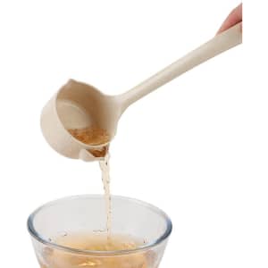 A wheat straw oil filter spoon in a light beige colour pouring liquid into a glass bowl. It features a logo.