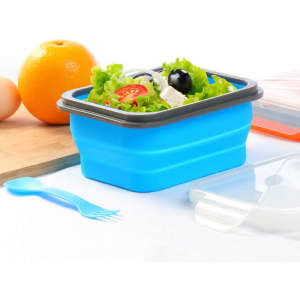 A blue collapsible lunch box with tableware, filled with salad and surrounded by fresh ingredients like an orange and eggs.