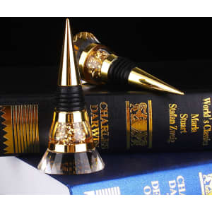 A drink bottle stopper in gold and clear crystal, featuring a sleek design and a black rubber base, rests on a stacked book.