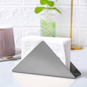 A stainless steel triangle napkin stand holding white napkins, set on a table with a glass and a plant in the background.