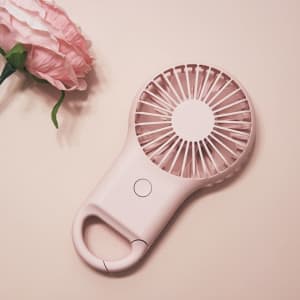 A pink mini rechargeable fan with a carabiner, placed beside a pink flower on a light background.