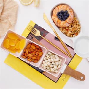 A white handheld snack serving bowl with compartments for almonds, pistachios, oranges, and granola, displayed with cutlery.