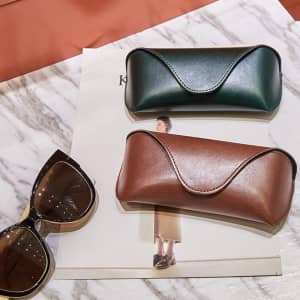 Two PU leather soft sunglasses cases, one dark green and one brown, placed on a marble surface with sunglasses nearby.