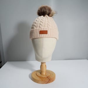 A cream cable-knit beanie featuring a fluffy fox fur pom pom, displayed on a wooden mannequin head.