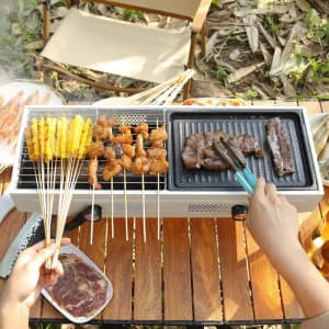 A multifunctional folding charcoal BBQ grill with grilled meats and skewers on a wooden table surrounded by greenery.