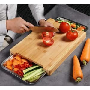 A bamboo chopping board featuring two stainless steel trays, with sliced vegetables and tomatoes on the surface.