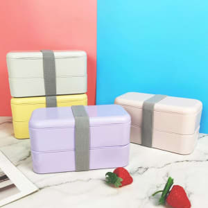 Double layer lunch boxes in pastel colours: grey, yellow, lavender, and pink, secured with grey straps on a marble surface.