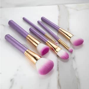 A set of six soft bristle makeup brushes with glittery purple handles and pink-tipped bristles, displayed on a marble surface.
