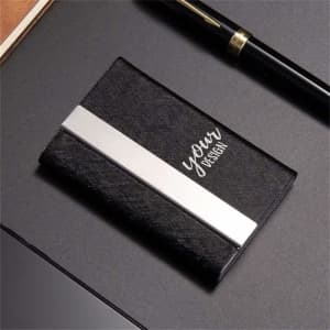 A black PU leather business card case with a silver stripe and a logo, resting on a dark surface beside a pen.