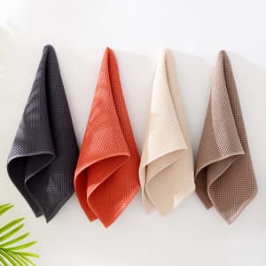 Four face towels in dark grey, terracotta, cream, and light brown, hanging on a wall with a plant partially visible.