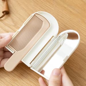 A foldable hand mirror in white with a tan-coloured comb, resting on a wooden surface.