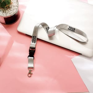 A silver custom lanyard with a buckle, featuring a logo, rests on a pink surface alongside a laptop and a plant.